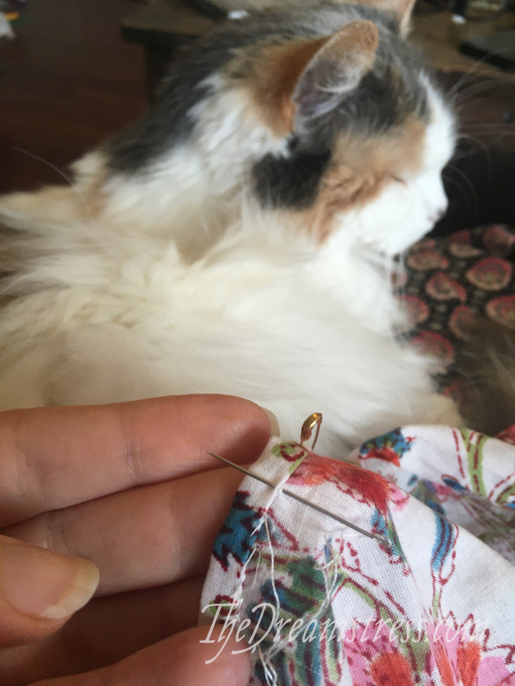 In the foreground, a hand sewing a narrow hem in floral chintz, In the background, a calico cat