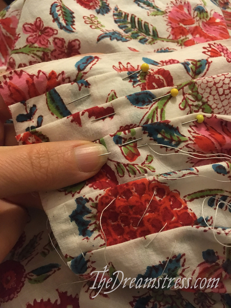 A hand holding a length of floral fabric with pleats secured with basting stitches