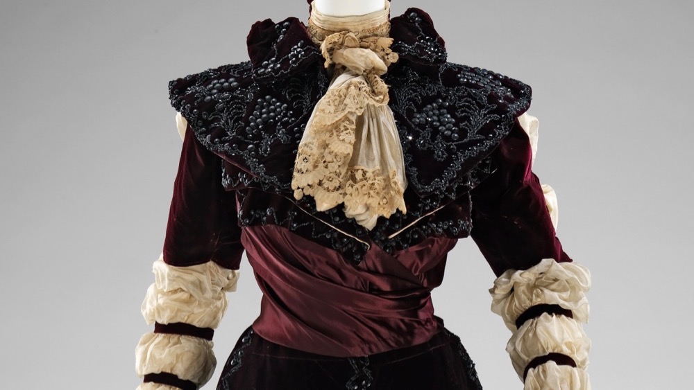 Image showing the front view of the bodice of an 1890s reception gown with sheer white sleeves gathered into 7 rows of puffs, a lace jabot, wine red satin bodice, and heavily beaded darkest red velvet skirt, collar and sleeves