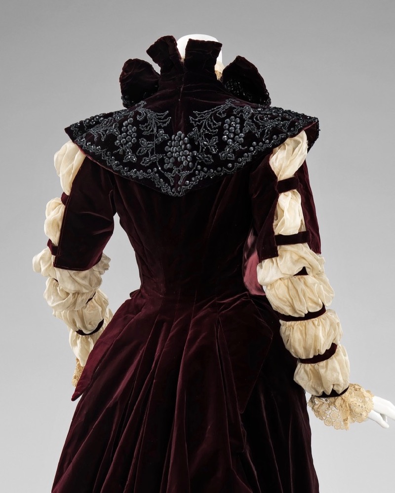 Image showing the back view of an 1890s reception gown with sheer white sleeves gathered into 7 rows of puffs, a standing collar, and a ruffle-edged train of darkest red velvet