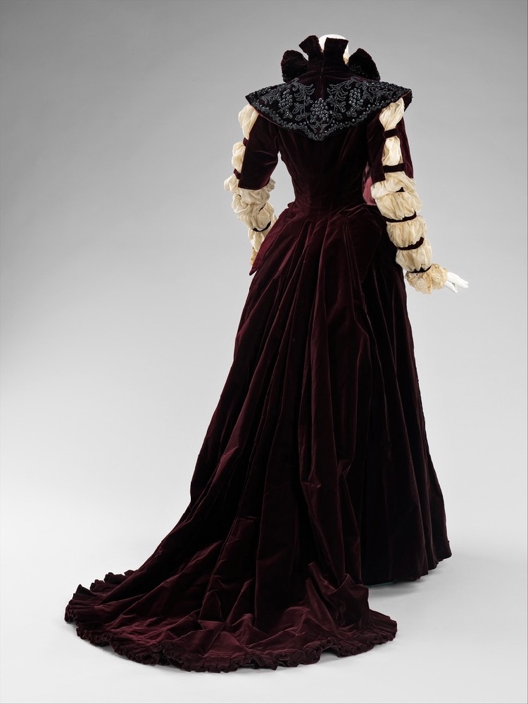 Image showing the back view of an 1890s reception gown with sheer white sleeves gathered into 7 rows of puffs, a standing collar, and a ruffle-edged train of darkest red velvet