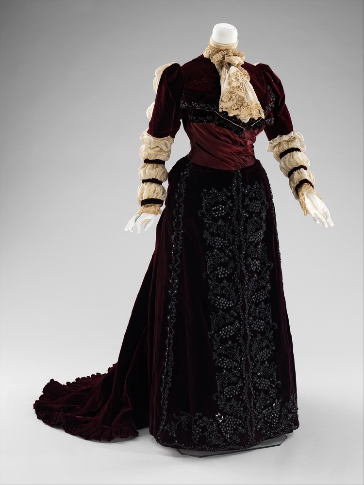 Image showing the front view of an 1890s reception gown with sheer white sleeves gathered into 7 rows of puffs, a lace jabot, wine red satin bodice, and heavily beaded darkest red velvet skirt, collar and sleeves