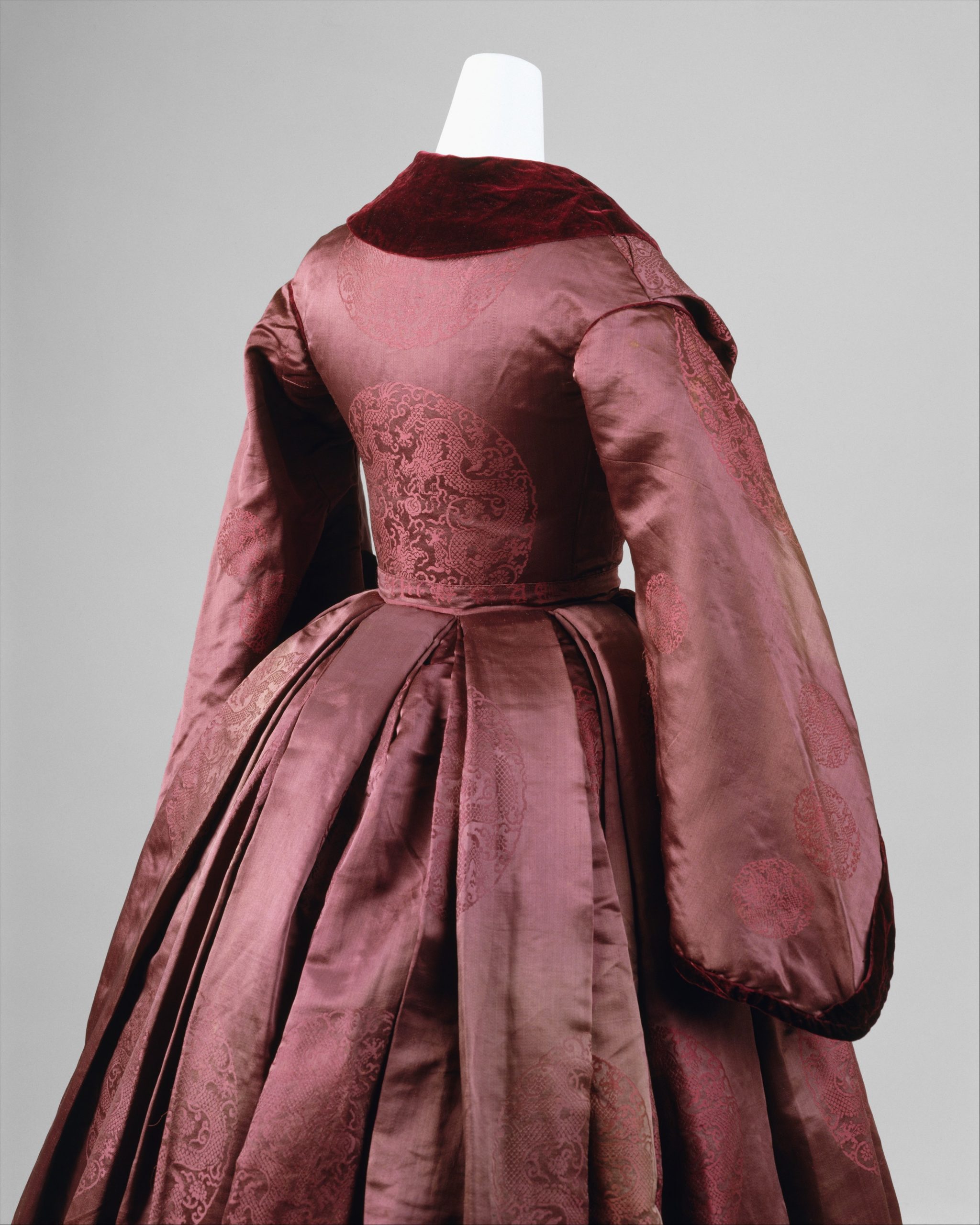 Image shows the upper half of a dark purple-brown bcoraded satin with velvet trimmings mid 19th century dress from the side-back
