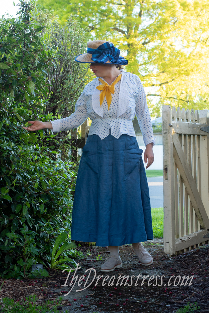 A woman in a mid-1910s outfit comprising a blue linen skirt with triangular pockets, and a polka dotted blouse stands in an open gate. She looks to her right, and touches a branch of a shrub