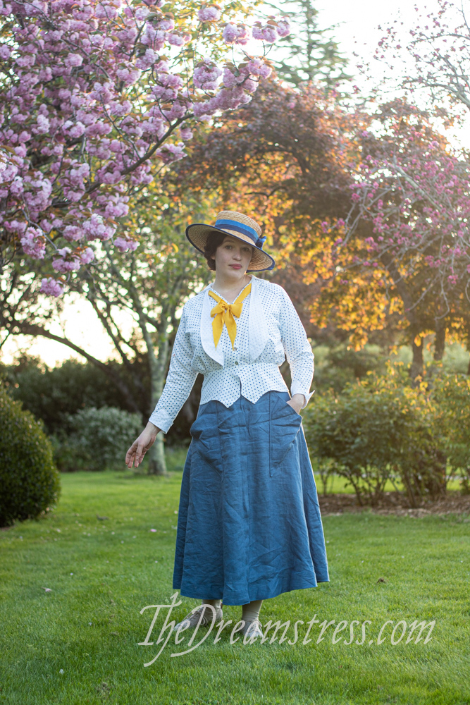 A woman in a mid-1910s outfit comprising a blue linen skirt with triangular pockets, and a polka dotted blouse stands underneath flowering cherry trees