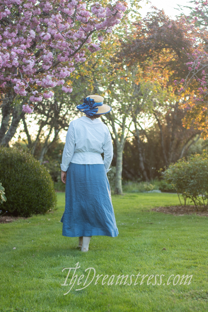 A woman in a mid-1910s outfit comprising a blue linen skirt with triangular pockets, and a polka dotted blouse walks away from the camera underneath flowering cherry trees