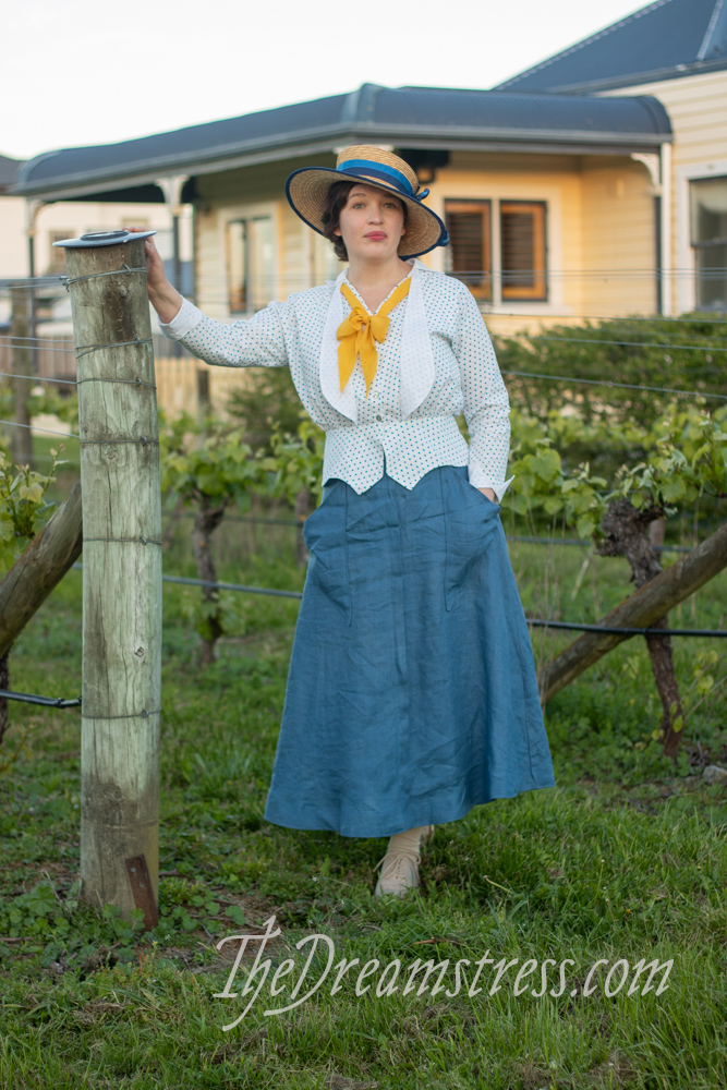 A woman in a mid-1910s outfit comprising a blue linen skirt with triangular pockets, and a polka dotted blouse stands in a vineyard with a house in the background