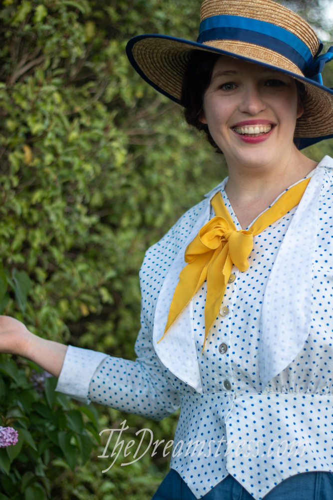A woman in a blue and white polka dotted 1910s blouse with yellow silk jabot, and broad brimmed straw hat trimmed with blue looks upward and smiles