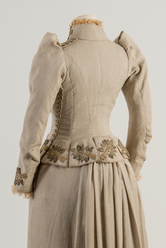 Image shows the back view of a light grey-beige jacket, very fitted, with full sleeves and leaf themed embroidery around the tail of the jacket.