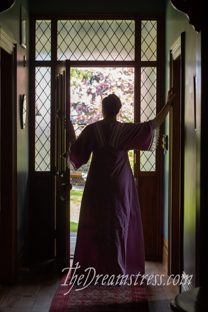 A woman in a long purple dress with wide sleeves stands silhouetted against a doorway, her arms raised