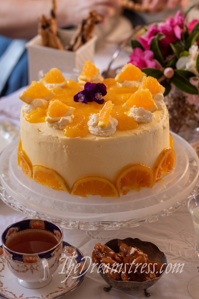 Image shows a round cake decorated with half circles of candied oranges, swirls of cream, slices of orange, and purple pansies