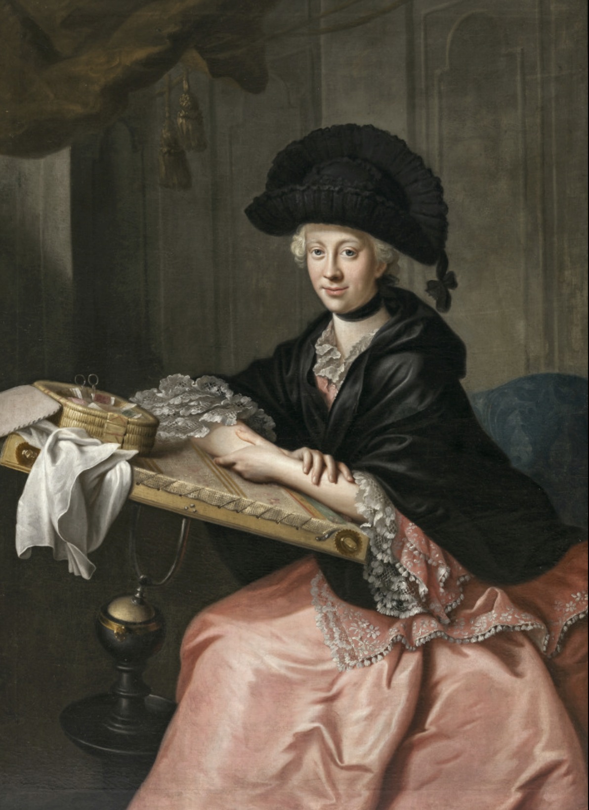 An 18th century portrait showing a lady with powdered hair, seated at a slanted table with her sewing basket in front of her, wearing a black silk mantle over a pink dress, with striking black hat