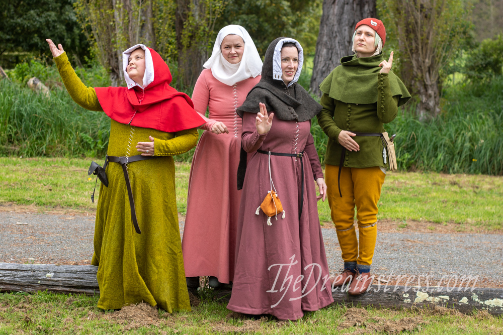 Four people in Medieval costume imitate poses from illuminated manuscripts. On the left a woman in a red hood and green-yellow gown raises her arm. Next to her a woman in a pink dress and white wimple holds her hands demurely in front of her. In front of her a woman in a pink-purple dress and grey hood gestures. On the far right a man in a green tunic and yellow leggings raises a hand.