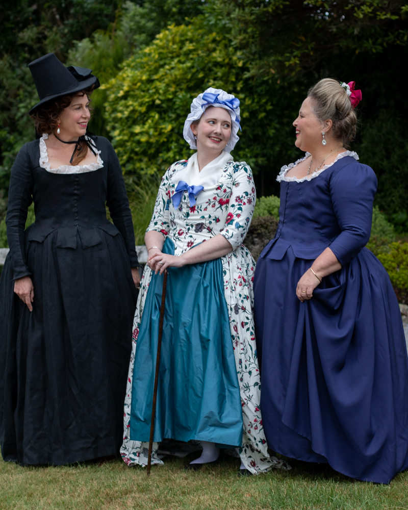 Three women in 18th century dress: a blond in cobalt blue, a woman in a cap in floral, and an woman with auburn curls in black, stand together. The woman in blue looks at the other two, who look back at her