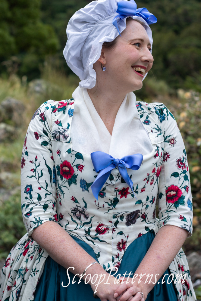 A fair skinned woman in a ruffly white cap topped with a bow stands in 3/4 view, her head turned over her left shoulder. She is wearing a late 18th century dress in bright florals on white with a fitted bodice with a blue bow at the bust, full skirt, elbow length sleeves, a white fichu filling in the neck, and a teal skirt. Her right hand picks up her floral overskirt, and her left is resting on a dark cane with a silver handle.