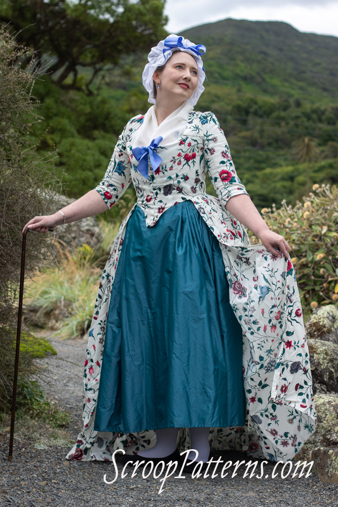A fair skinned woman in a ruffly white cap topped with a bow stands in 3/4 view, her head turned over her left shoulder. She is wearing a late 18th century dress in bright florals on white with a fitted bodice with a blue bow at the bust, full skirt, elbow length sleeves, a white fichu filling in the neck, and a teal skirt. Her right hand picks up her floral overskirt, and her left is resting on a dark cane with a silver handle.