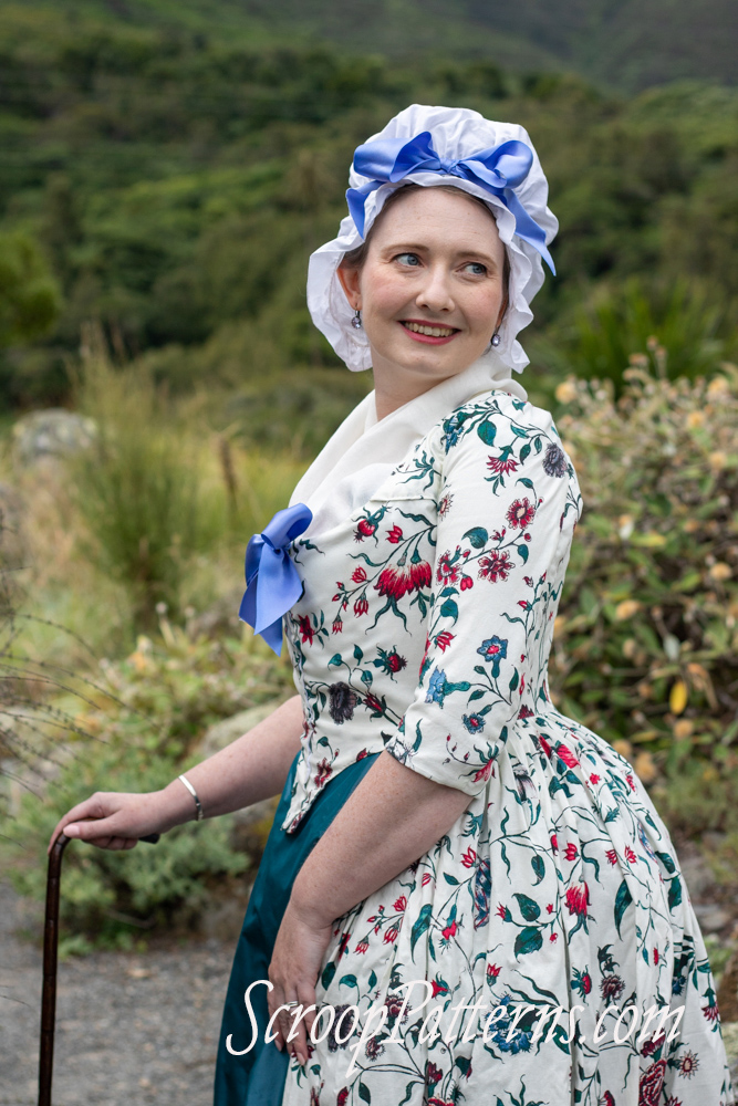 A fair skinned woman in a ruffly white cap topped with a bow stands in profile, and turns her head over her shoulder to look back. She is wearing a late 18th century dress in bright florals on white with a fitted bodice with a blue bow at the bust, full skirt, elbow length sleeves, a white fichu filling in the neck, and a teal skirt. She is resting on a dark cane with a silver handle.