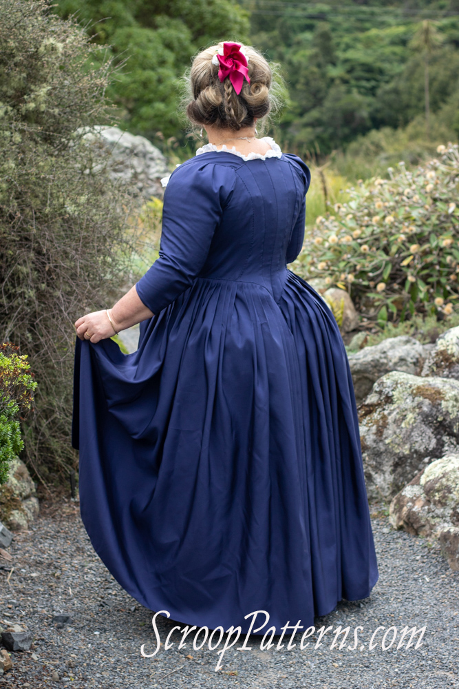 A woman with blond hair styled on top of her head stand with her side back to us. She is wearing a late 18th century dress in cobalt blue silk. Her left hand picks up her skirts to show the fall of the pleating. 