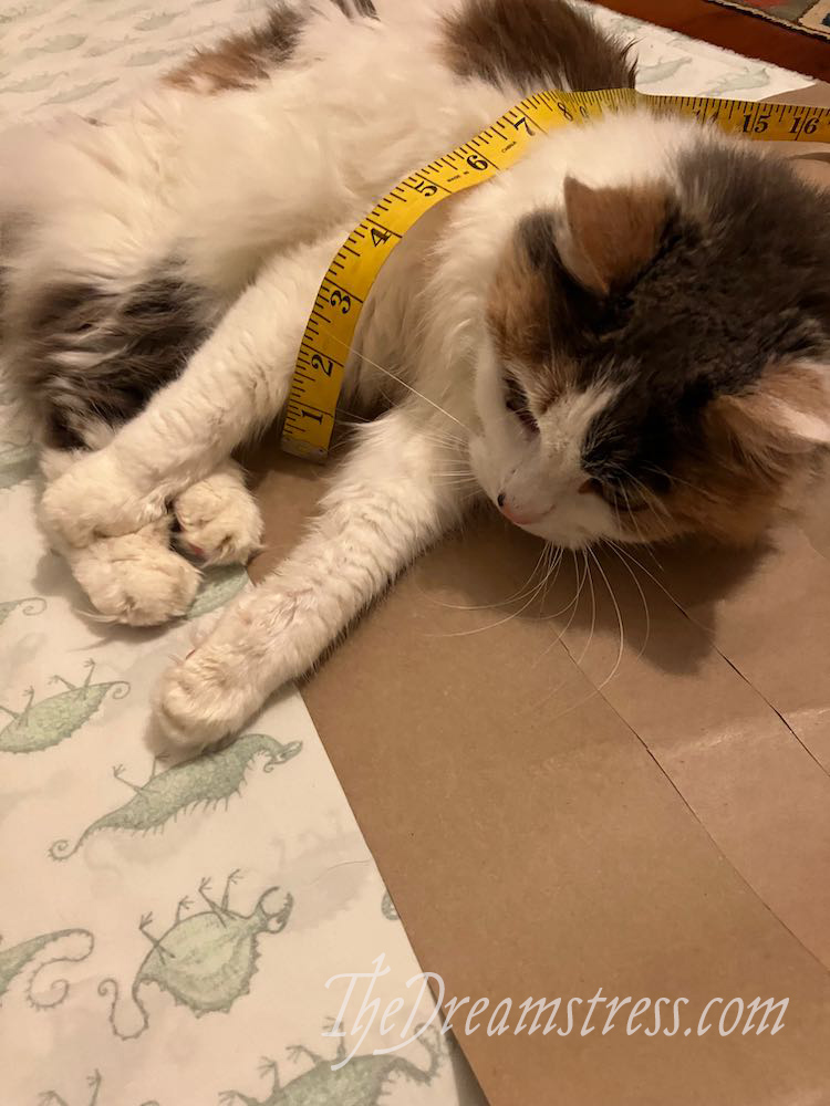 A calico cat lies on brown pattern paper and white fabric with green monsters, a yellow measuring tape wrapped around her.