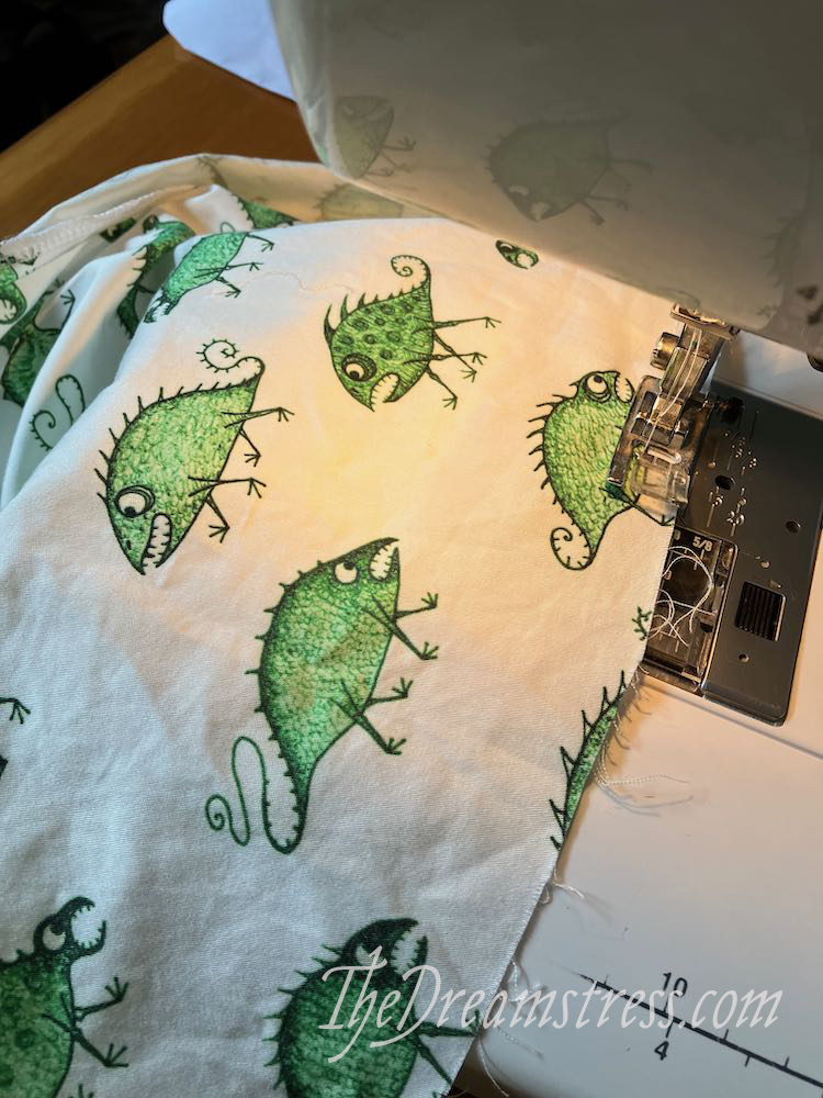 Image shows a sewing machine stitching white fabric with a pattern of green monsters